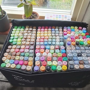 Puede incluir: Un estuche negro con cremallera lleno de una gran colección de rotuladores de arte coloridos. Los rotuladores están dispuestos en filas, con una variedad de colores visibles, incluyendo tonos de verde, rosa, azul y amarillo. El estuche está abierto.