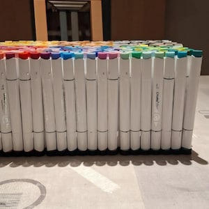 May include: A large collection of white-barreled markers arranged in a dense, rectangular block. Each marker features a colored cap corresponding to its ink shade, creating a vibrant spectrum of colors across the top of the organized set.