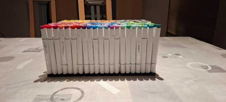 May include: A large collection of white-barreled markers arranged in a dense, rectangular block. Each marker features a colored cap corresponding to its ink shade, creating a vibrant spectrum of colors across the top of the organized set.