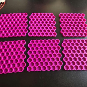 May include: Six square coasters in a vibrant magenta pink color, each featuring a repeating honeycomb pattern of circular cutouts. The coasters are arranged in two rows of three, displaying a uniform grid structure with rounded edges.