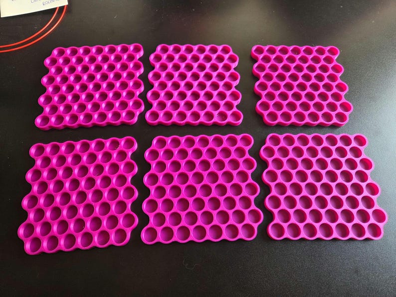 May include: Six square coasters in a vibrant magenta pink color, each featuring a repeating honeycomb pattern of circular cutouts. The coasters are arranged in two rows of three, displaying a uniform grid structure with rounded edges.