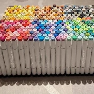 May include: A large rectangular collection of white-bodied markers arranged vertically in a dense grid. The marker caps display a wide spectrum of colors, including shades of red, pink, orange, yellow, blue, purple, green, brown, gray, and black.