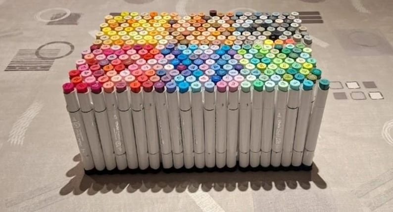 May include: A large rectangular collection of white-bodied markers arranged vertically in a dense grid. The marker caps display a wide spectrum of colors, including shades of red, pink, orange, yellow, blue, purple, green, brown, gray, and black.