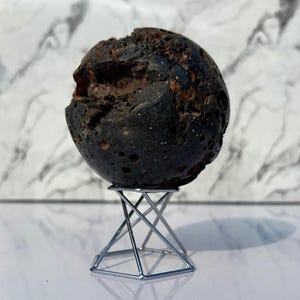 Puede incluir: Una piedra oscura y esférica con una superficie rugosa y texturizada, exhibida sobre un soporte de metal plateado. La piedra tiene un exterior gris oscuro con un interior marrón rojizo visible en una cavidad. El soporte tiene un diseño geométrico en forma de pirámide.