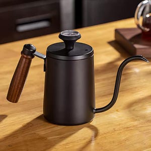 May include: A black gooseneck kettle with a wooden handle and a thermometer on top. The kettle is made of stainless steel and is designed for pour-over coffee brewing. The handle is made of dark brown wood. The kettle is sitting on a wooden surface.
