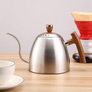 Può includere: Un bollitore a collo di cigno in acciaio inossidabile con manico e pomello del coperchio in legno. Il bollitore ha un lungo beccuccio curvo per versare con precisione. Un versatore di caffè rosso e una tazza bianca sono sullo sfondo.