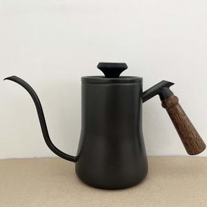 May include: Black gooseneck kettle with a wooden handle and lid knob. The kettle has a long, curved spout for precise pouring. The handle is made of wood with a natural grain pattern. The kettle is designed for brewing coffee or tea.