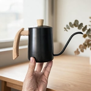 May include: A black gooseneck kettle with a wooden handle and lid knob. The kettle has a long, curved spout for precise pouring. The handle is made of light-colored wood, contrasting with the black body. A stylish coffee brewing accessory.