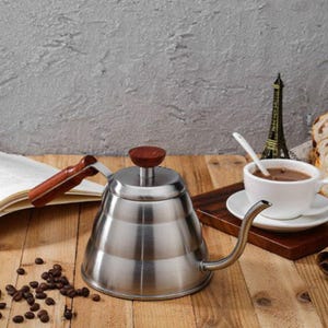 Può includere: Una caffettiera in acciaio inossidabile con manico e pomello in legno. La caffettiera è su una superficie di legno con chicchi di caffè, una tazza di caffè e un libro. Una piccola statua della Torre Eiffel è sullo sfondo.