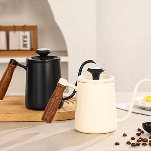 Può includere: Due bollitori per caffè versato. Uno è nero, l'altro color crema. Entrambi hanno un beccuccio lungo e curvo, manici in legno e coperchi neri. I bollitori sono progettati per una preparazione precisa del caffè.