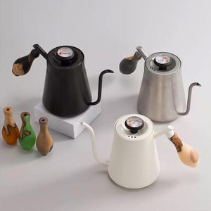 May include: Three gooseneck kettles in black, silver, and white, each with a wooden handle and a built-in thermometer. Small decorative wooden vases in shades of orange, green, and brown are in the foreground. The kettles are designed for pour-over coffee.