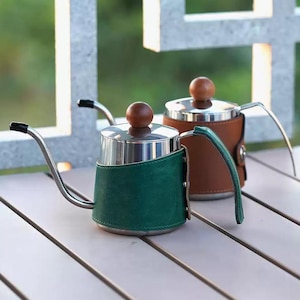May include: Two stainless steel gooseneck kettles with wooden knobs and leather wraps. One kettle has a green leather wrap, the other a brown one. The kettles have long, thin spouts and are designed for precise pouring.