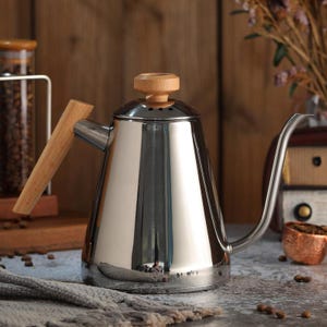 Könnte beinhalten: Eine polierte Edelstahl-Schwanenhals-Kanne mit Holzgriff und Deckelknopf. Der lange, gebogene Ausguss ermöglicht präzises Ausgießen. Modernes Design, ideal für Pour-Over-Kaffee.