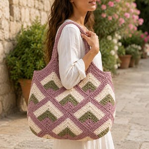 Puede incluir: Un bolso tote de ganchillo grande con un patrón geométrico de chevrón en malva, crema y verde oliva. El bolso tiene dos correas anchas para el hombro y está construido con motivos cuadrados interconectados, creando un diseño triangular texturizado.