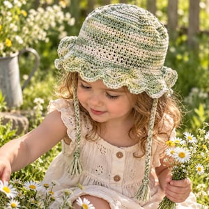 May include: A crochet bucket hat featuring a variegated color palette of sage green, cream, and soft yellow. The hat has a wide, ruffled brim and two braided chin straps that hang down, each finished with a small tassel at the bottom end.
