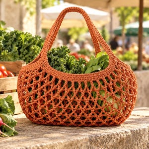 May include: A burnt orange crochet market bag with an open diamond mesh pattern and two integrated handles, filled with fresh green kale and leafy vegetables, sitting on a stone surface next to red radishes and tomatoes.