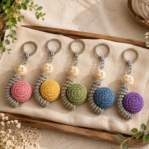 May include: Five handmade crochet snail keychains with silver metal rings. Each snail features a light gray crocheted body, a round wooden bead head with a painted smiling face, and a spiral shell in pink, yellow, green, blue, or purple.