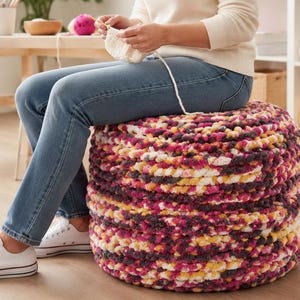 May include: A round, floor-sitting ottoman featuring a thick, chunky knit texture. The yarn is a variegated blend of vibrant magenta, deep charcoal gray, bright yellow, and cream white, creating a dense, multi-colored, soft-touch surface.