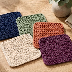 May include: A set of five square coasters featuring a thick, braided crochet texture. The coasters are available in solid colours including navy blue, sage green, burnt orange, cream, and deep purple, each with a consistent woven pattern.