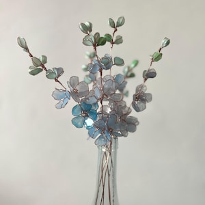 May include: A decorative arrangement of handcrafted flowers made from translucent light blue and green resin petals with thin copper wire stems. The flowers are displayed in a clear glass bottle, featuring delicate, multi-layered petals.