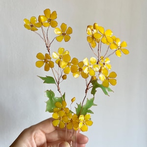 Może przedstawiać: Ręcznie wykonana kompozycja kwiatowa z jasnożółtymi kwiatami o pięciu płatkach, zielonymi liśćmi i łodygami z drutu miedzianego. Kwiaty są prezentowane w małym, przezroczystym szklanym wazonie. To element dekoracyjny.