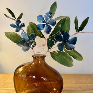 May include: A decorative floral arrangement with blue metal flowers and green leaves, displayed in a brown glass vase. The arrangement is made from metal and wire. The vase is placed on a wooden surface, creating a unique artistic display.