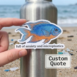 May include: A sticker featuring a blue and orange fish with the text "full of anxiety and microplastics." A second sticker with the text "Custom Quote" is also visible. The stickers are held against a silver water bottle on a sandy beach.