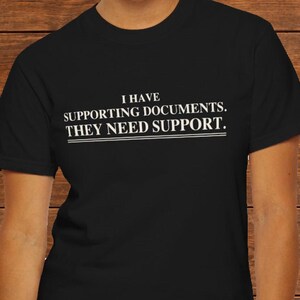 May include: A black short-sleeved t-shirt featuring white text printed on the chest that reads: I HAVE SUPPORTING DOCUMENTS. THEY NEED SUPPORT. The text is arranged in three lines with a double horizontal line underneath the final sentence.