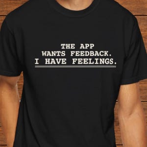 May include: A black short-sleeved t-shirt featuring white text printed on the chest that reads: THE APP WANTS FEEDBACK. I HAVE FEELINGS. The shirt has a crew neckline and a standard fit, with the text centered in a bold, sans-serif font.