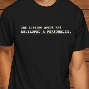 May include: A black short-sleeved t-shirt featuring white text printed on the chest that reads: THE EDITING QUEUE HAS DEVELOPED A PERSONALITY. The text is written in a typewriter-style font and is underlined with a solid white line.