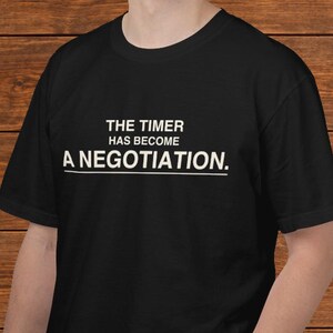 May include: A black short-sleeved t-shirt featuring white text printed on the chest that reads THE TIMER HAS BECOME A NEGOTIATION. The lettering is arranged in three lines with a horizontal line underneath the final word.