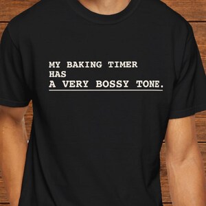 May include: A black short-sleeved t-shirt featuring white text that reads: MY BAKING TIMER HAS A VERY BOSSY TONE. The lettering is printed in a typewriter-style font with a horizontal line underneath the text centered on the chest area.