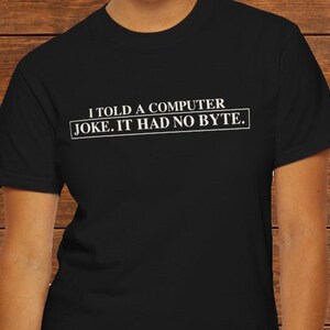 May include: A black short-sleeved t-shirt featuring white text printed on the chest that reads: I TOLD A COMPUTER JOKE. IT HAD NO BYTE. The text is arranged in two lines and enclosed within a thin white rectangular border.