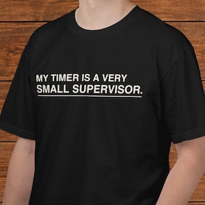 May include: A black short-sleeved t-shirt featuring the white text "MY TIMER IS A VERY SMALL SUPERVISOR." printed on the chest. The text is arranged in two lines with a horizontal line underneath the words, creating a simple graphic design.