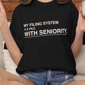 May include: A black short-sleeved t-shirt featuring white text that reads: MY FILING SYSTEM IS A PILE WITH SENIORITY. The shirt has a crew neckline and a standard fit, displayed as a plain garment without any additional patterns or graphics.