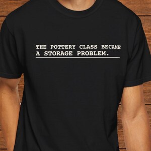 May include: A black short-sleeved t-shirt featuring white text printed on the chest that reads, THE POTTERY CLASS BECAME A STORAGE PROBLEM, with a thin horizontal white line positioned directly underneath the text.