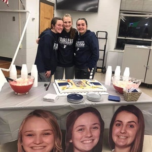 May include: Four young women stand in front of a table with a cake decorated with a basketball court design. The cake is surrounded by blue and gray plates. The women are wearing blue sweatshirts with the word "Fairless" on them. The table is covered with a gray tablecloth and there are two pitchers of punch on the table.