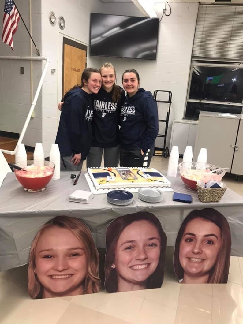 May include: Four young women stand in front of a table with a cake decorated with a basketball court design. The cake is surrounded by blue and gray plates. The women are wearing blue sweatshirts with the word "Fairless" on them. The table is covered with a gray tablecloth and there are two pitchers of punch on the table.