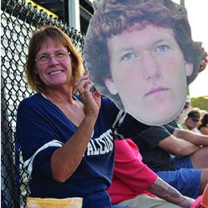 May include: A woman wearing a blue shirt and glasses is holding a cardboard cutout of a man's face. The cutout is large and covers most of the woman's body. The woman is smiling and looking at the camera. The cutout is of a man with brown hair and blue eyes. He is wearing a white shirt. The woman is sitting in a bleacher with a bag of chips in her lap.