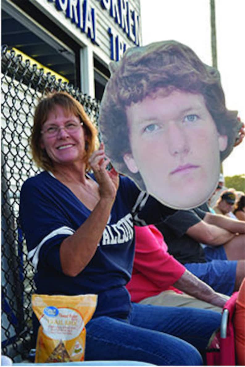May include: A woman wearing a blue shirt and glasses is holding a cardboard cutout of a man's face. The cutout is large and covers most of the woman's body. The woman is smiling and looking at the camera. The cutout is of a man with brown hair and blue eyes. He is wearing a white shirt. The woman is sitting in a bleacher with a bag of chips in her lap.