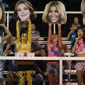 Puede incluir: Cuatro personas sostienen grandes recortes de cart&oacute;n de caras. Las personas est&aacute;n sentadas en las gradas de un evento deportivo. Llevan collares de flores y sonr&iacute;en.