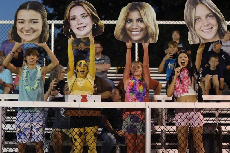 Puede incluir: Cuatro personas sostienen grandes recortes de cart&oacute;n de caras. Las personas est&aacute;n sentadas en las gradas de un evento deportivo. Llevan collares de flores y sonr&iacute;en.