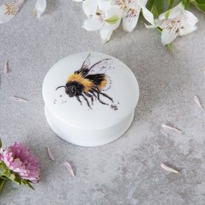 May include: A white ceramic trinket box with a round lid featuring a hand-painted illustration of a bumblebee with yellow and black stripes and brown eyes. The bumblebee is flying with its wings spread.