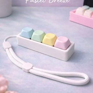 May include: A white rectangular accessory with four pastel-colored keycaps: blue, green, yellow, and pink. A white wrist strap is attached. The text "Pastel Breeze" is above the accessory.