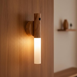 Puede incluir: Una lámpara de pared de madera con una luminaria cilíndrica. La lámpara está hecha de madera de color claro y tiene una base circular. La luminaria es de color blanco esmerilado, emitiendo un brillo suave. La lámpara está montada en una pared de madera.