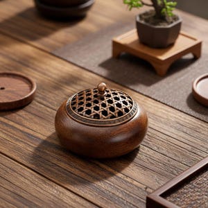 Op de afbeelding: Een houten wierookbrander met een decoratief bronskleurig bovenstuk met een geweven patroon. De brander is rond met een kleine bronzen knop bovenop. De achtergrond bevat andere houten voorwerpen, wat wijst op een thema van ontspanning en interieurdecoratie.