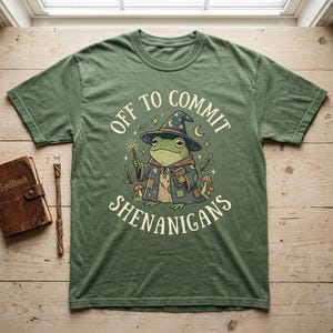 May include: Green t-shirt with the phrase "OFF TO COMMIT SHENANIGANS" and a cartoon frog wearing a wizard hat and robe. The frog is holding a wand and a book. A spellbook and wand are on the left.