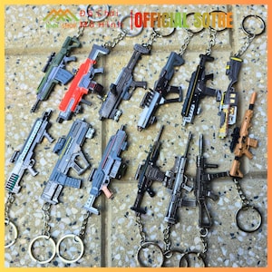 Può includere: Una collezione di portachiavi a forma di pistola in vari colori, tra cui verde, rosso, grigio e marrone. Ogni portachiavi è attaccato a una catena e un anello argentati. L'immagine contiene anche il testo "OFFICIAL SOTRE".