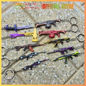May include: A collection of miniature weapon keychains in various designs and colors, including rifles, a dagger, and an axe. The keychains are attached to silver keyrings and are displayed on a textured surface. The text "OFFICIAL SOTRE" is visible.