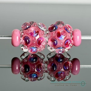 May include: Two spherical glass beads featuring a light pink base with raised, translucent circular accents in shades of blue, purple, and pink. The beads are strung on a thin metal rod, with a solid pink cylindrical bead on each end.